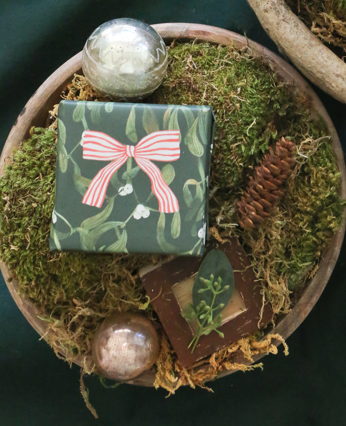 Gift wrapped with mistletoe and red bow wrapping paper with ornaments and a pine cone besides it 